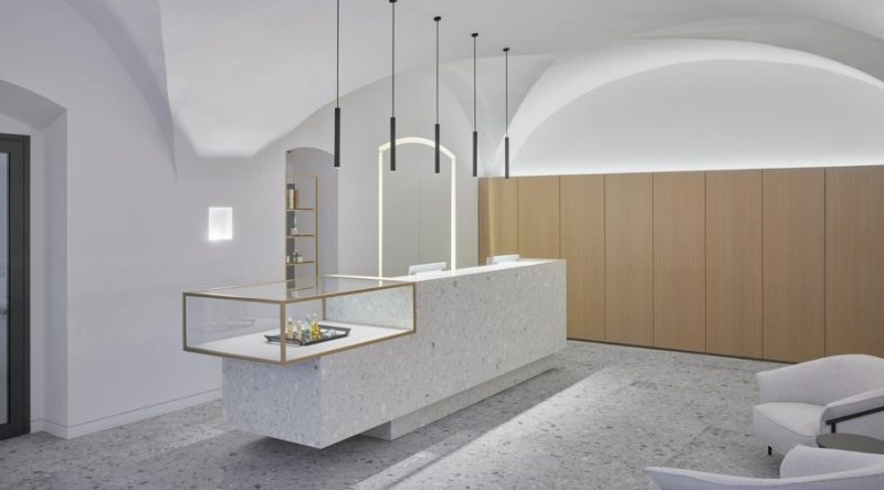 Minimalistinio stiliaus registratūros zona su ilga, šviesiai pilkos spalvos teraco registratūros stalą, stikline vitrina ir pakabinamais šviestuvais, kontrastuojanti su medinėmis sienų panelėmis ir skliautiniais lubais.